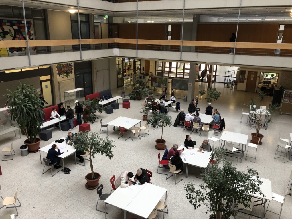 Das Foto zeigt den Eingangsbereich der Max-Weber-Schule im Erdgeschoss. Wenn viele Senioren:innen zur Smartphone-Sprechstunde kommen, dann beraten und helfen die Schülerinnen gerne auch hier. Auf dem Foto sind Tische, Stühle, Senior:innen und die Schüler:innen zu sehen.