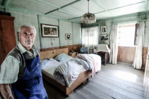 Das Bild zeigt einen alten Mann, der einsam wirkt. Er steht in seinem Schlafzimmer.